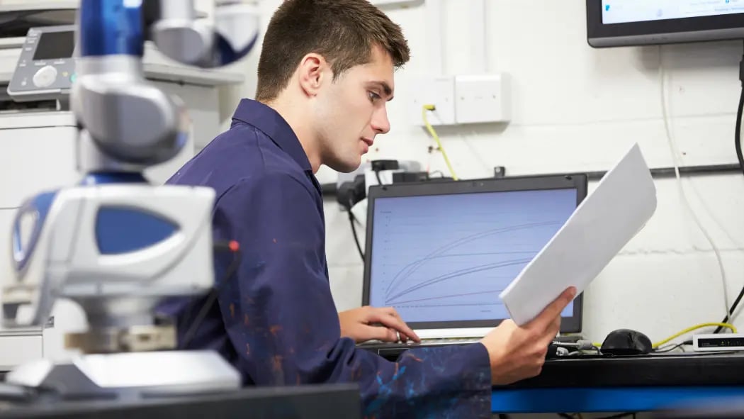 A man sat at his laptop at work reading a paper document