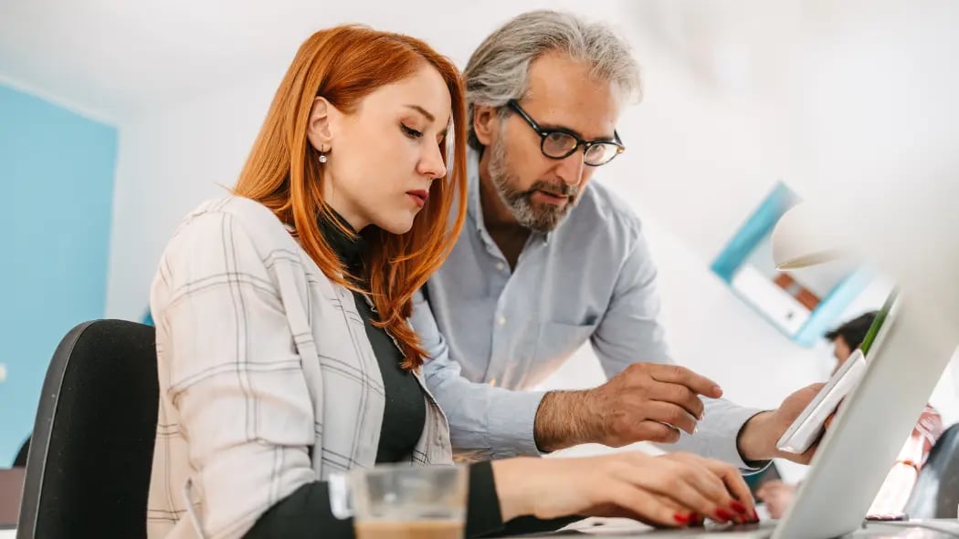 Woman sitting at desk on laptop with man standing behind pointing at the screen