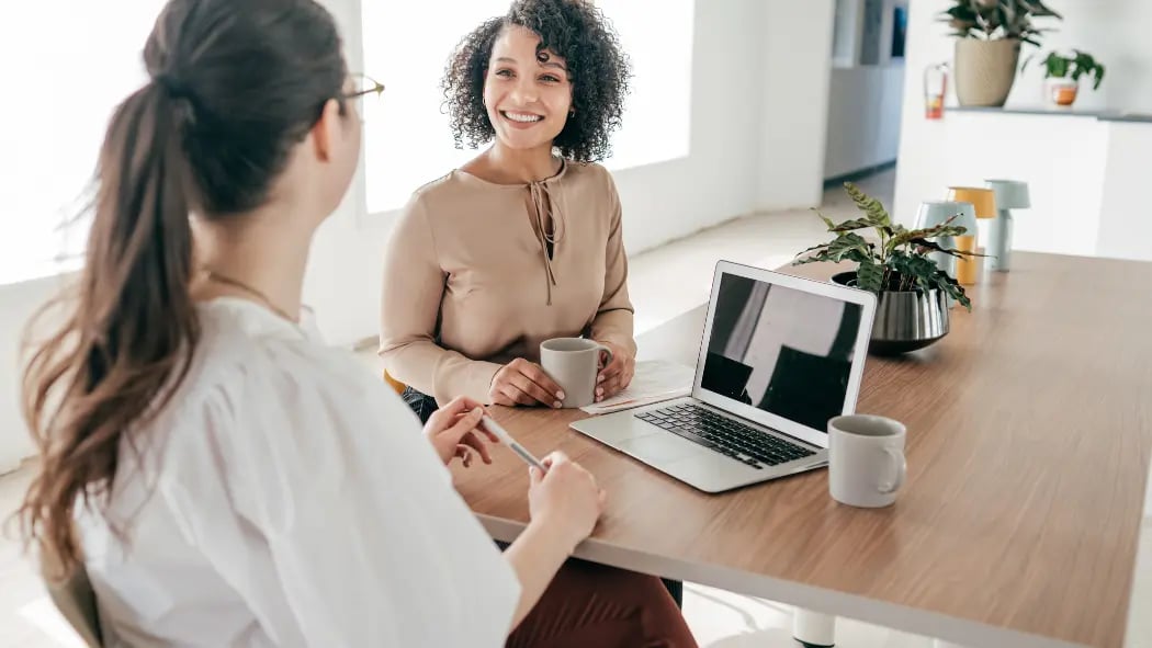Two women sat at desk talking with a laptop in between them