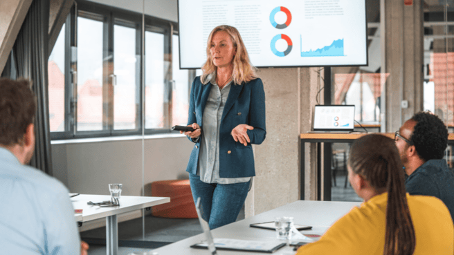 a woman standing at the front of a room of people presenting data