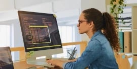 A young woman sitting at a desk looking at rows of data on a computer screen