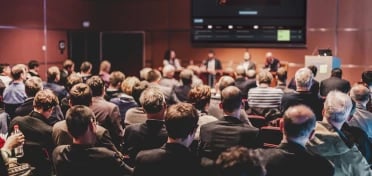 Conference with audience sat in front of a panel on stage with a screen above showing EDUKATE.AI learning platform