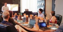 People sitting in a meeting room at a conference table while a man stands and gives a presentation at the front of the room