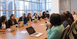 Ten or more people sitting around a long conference table in an office with laptops in front of them