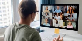 A man sat at a desk with a virtual meeting with several other people on a computer screen