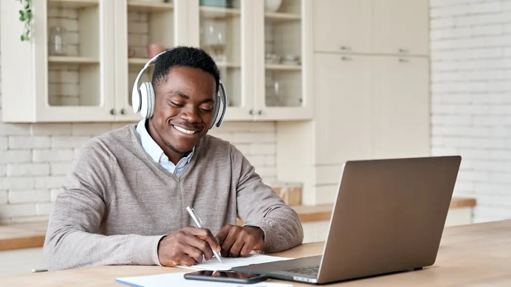A man sat at a laptop wearing headphones smiling an writing