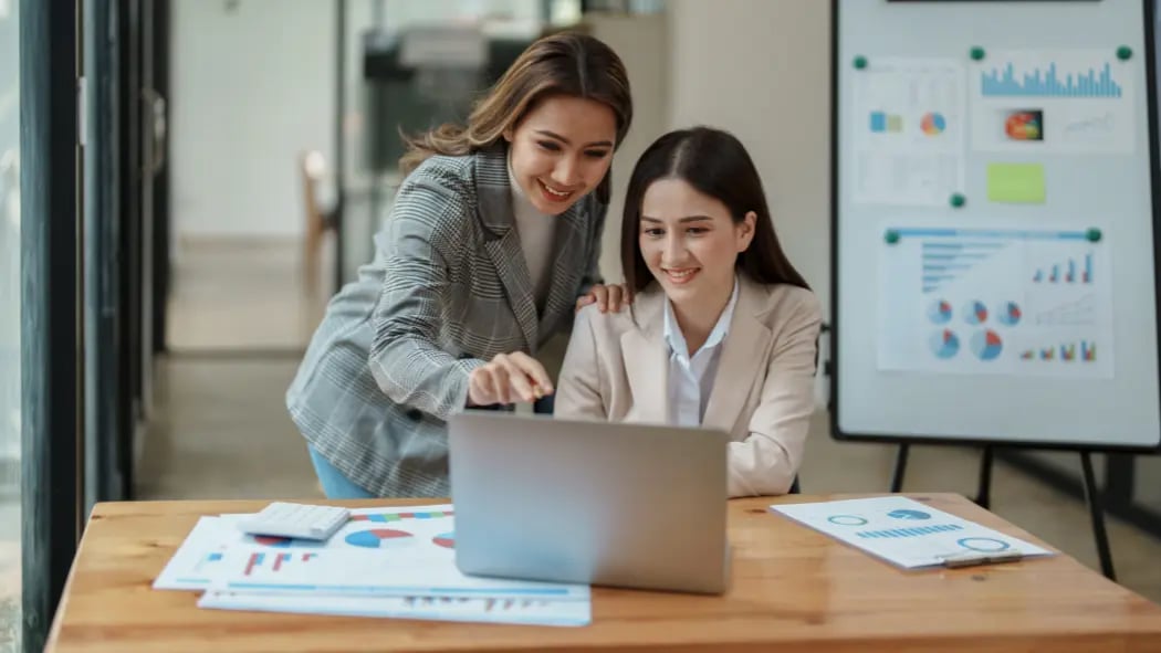 two women in office looking at computer screen with data on pieces of paper around them